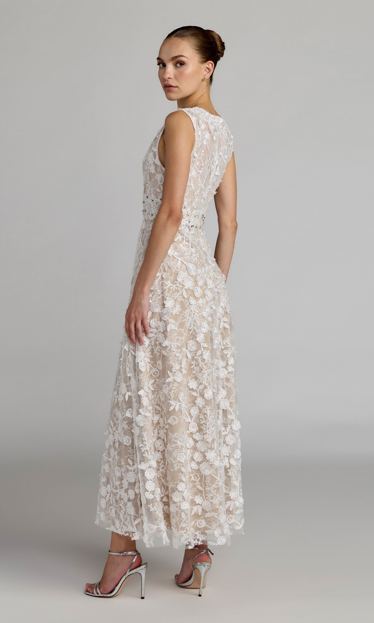 A woman stands in profile against a plain gray background, wearing the Mac Duggal 69192 tea-length party dress—an elegant sleeveless lace design with an A-line skirt—paired with silver high-heeled sandals.
